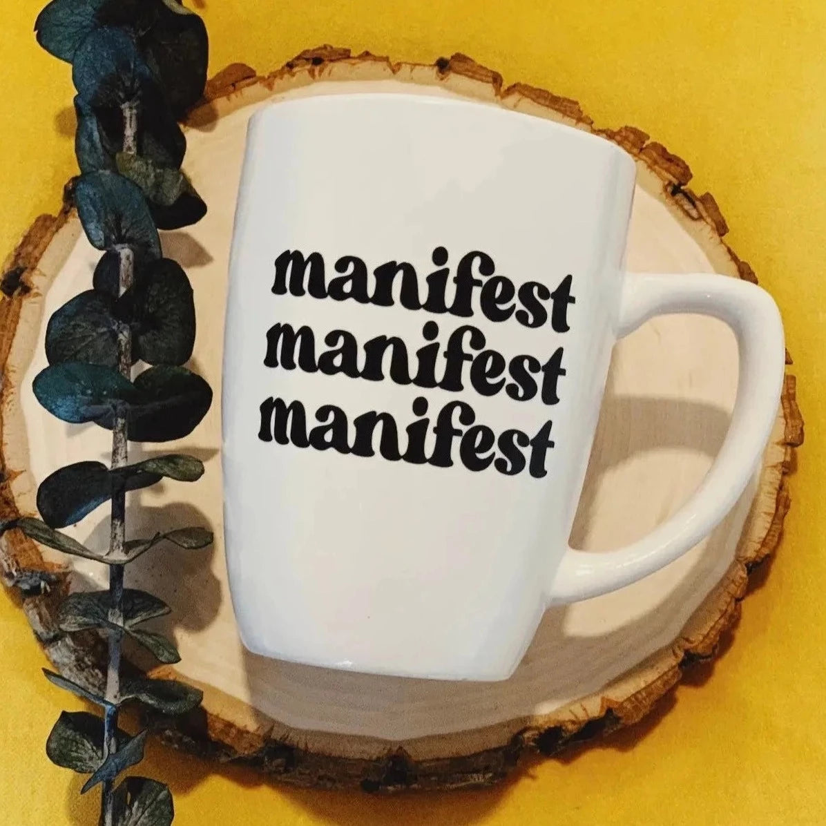 White mug with manifest manifest manifest in black on the front. On a yellow background, cut slab of wood with a piece of euceliptus  